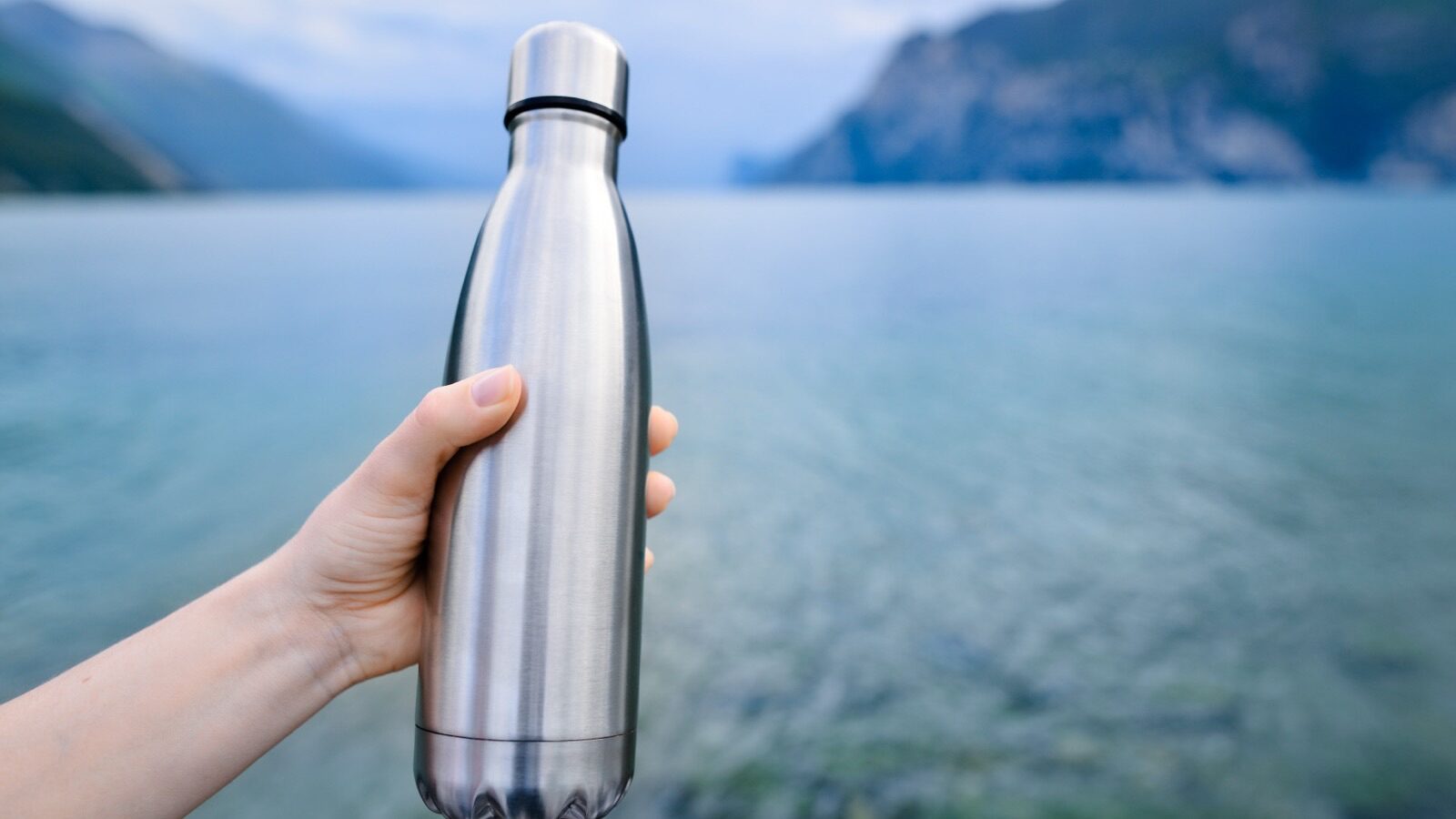 Main tenant une gourde isotherme en acier brossé devant un lac de montagne
